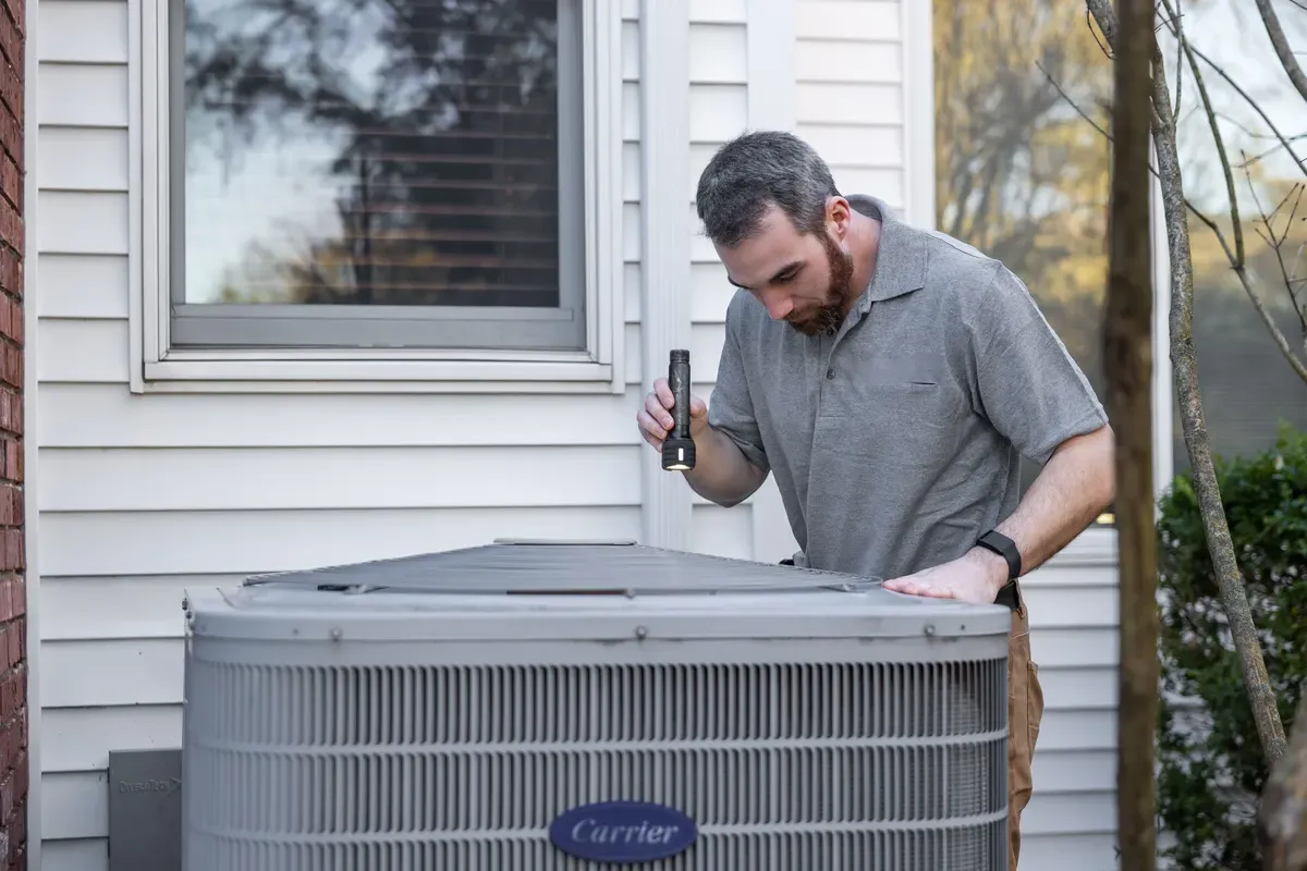 A technician inspecting a residential outdoor AC condenser with a flashlight